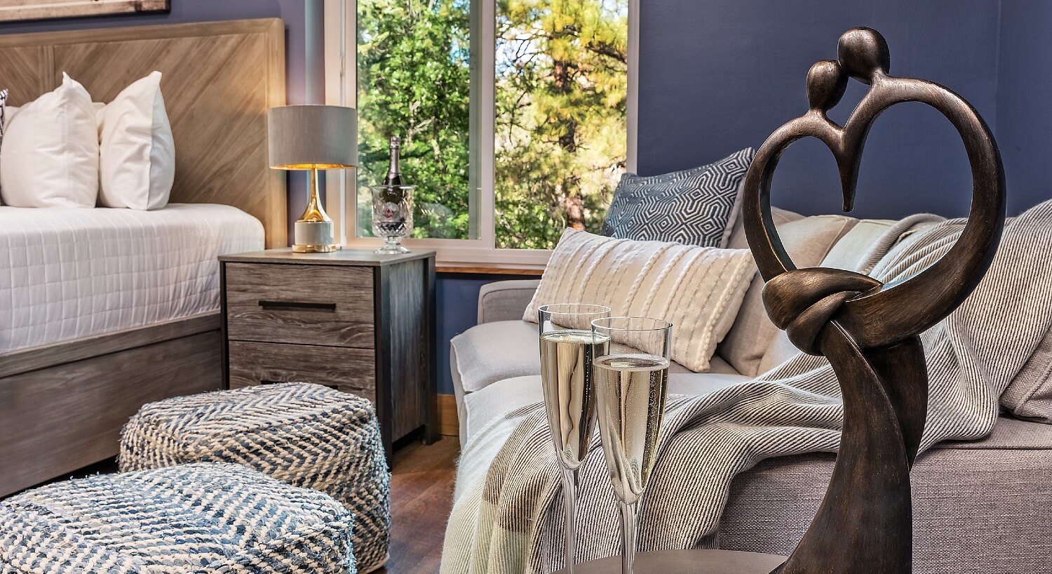 Corner of a luxurious guest room with bright window, grey loveseat and side table with two champagne glasses and metal art sculpture