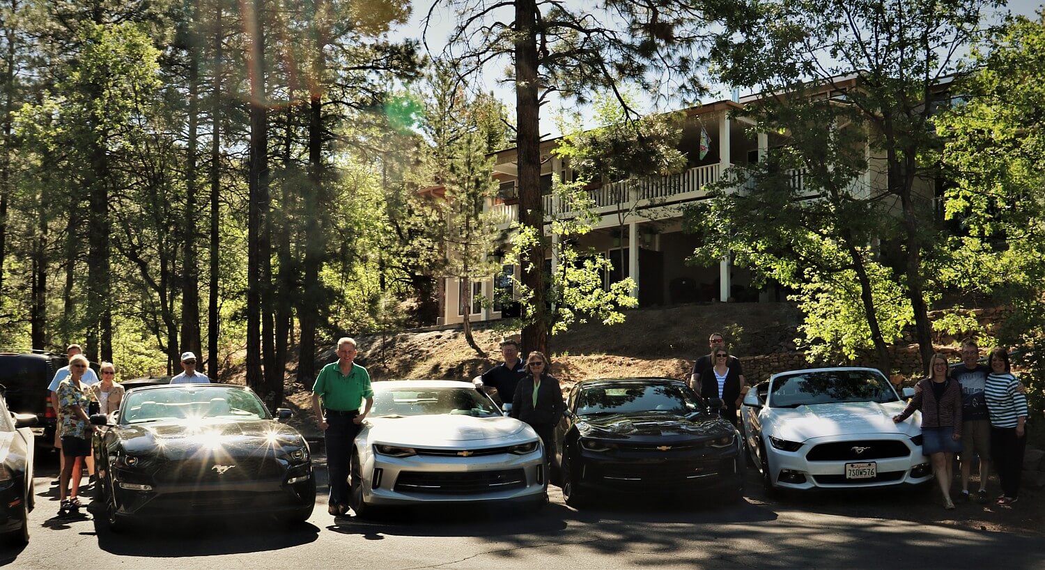 Parking lot with several people standing by black and white cars with trees and a large home up on a hill behind them