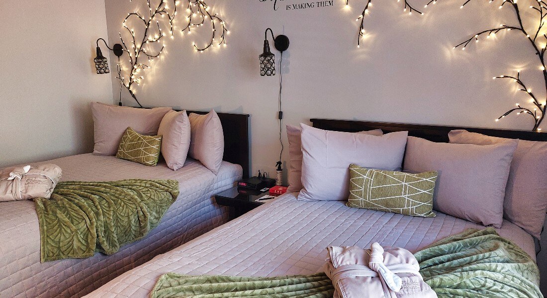 2 beds with gray comforters and green blankets with a robe on them and 4 pillows up against a lighted willow tree on the wall