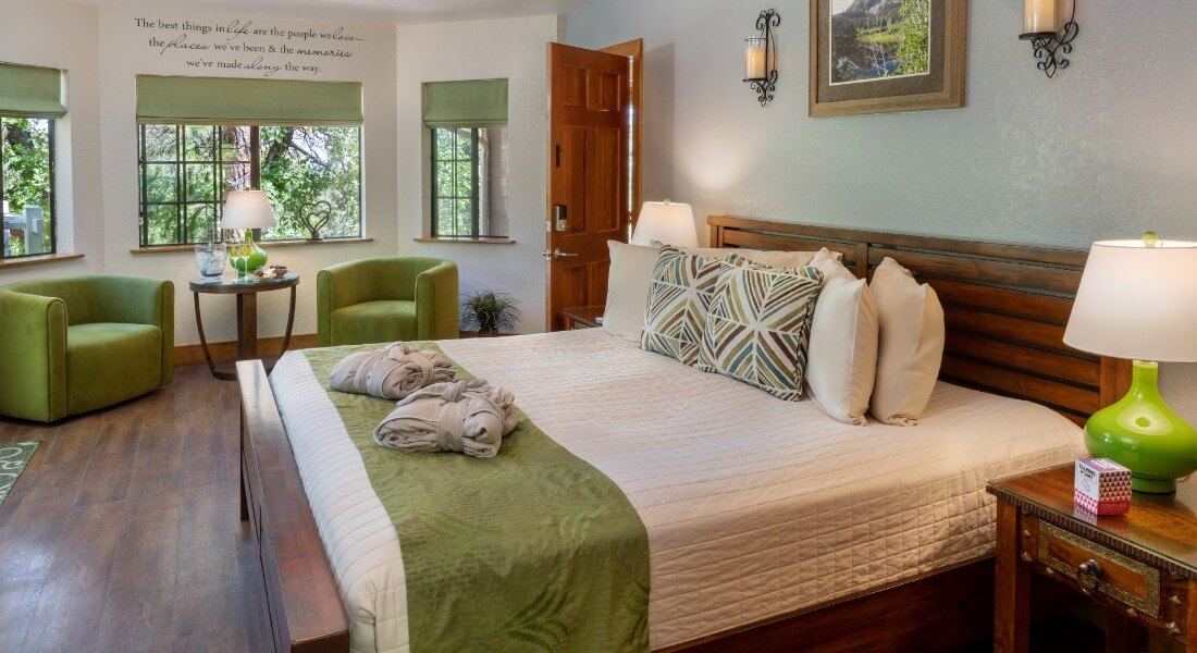 Luxurious king bed with white and green linens in a spacious bedroom with large windows and sitting area