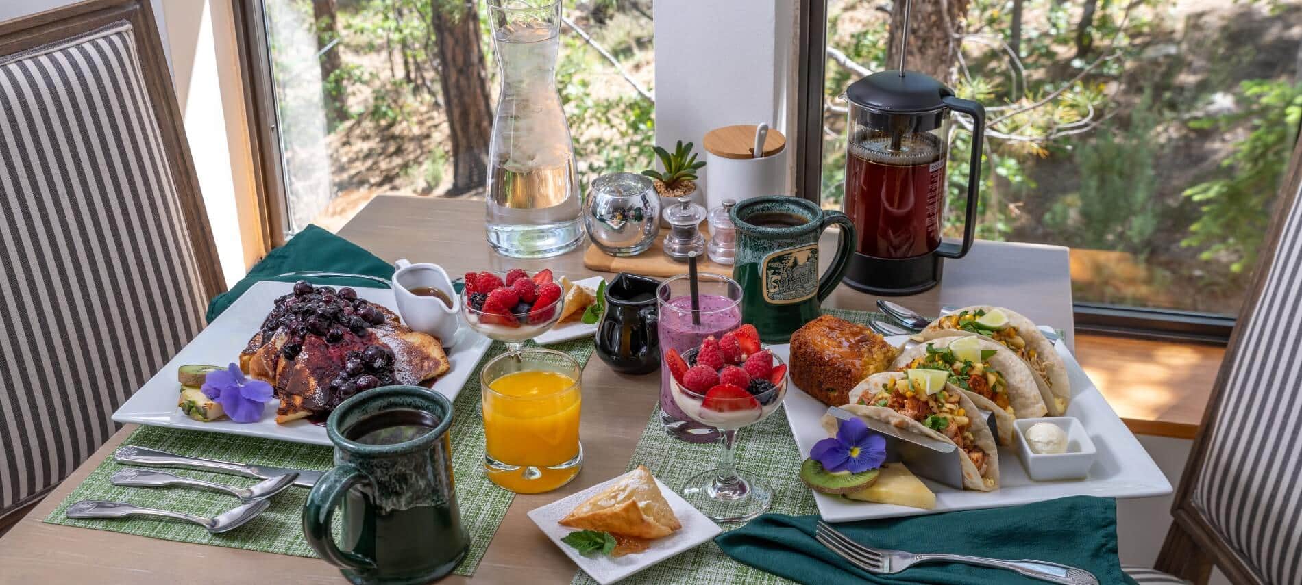 Cozy breakfast setup for two at the Sheridan House Inn, with beautifully plated entrees, fresh-squeezed orange juice, fruit smoothies, yogurt parfaits, branded coffee mugs, and a French press ready to be plunged.