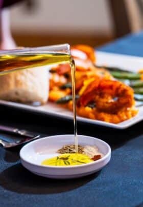 A stream of olive oil pours into a herb-filled dipping dish, with a dinner plate in the background showcasing a shrimp entrée served over sweet potato mash with asparagus and fresh bread.