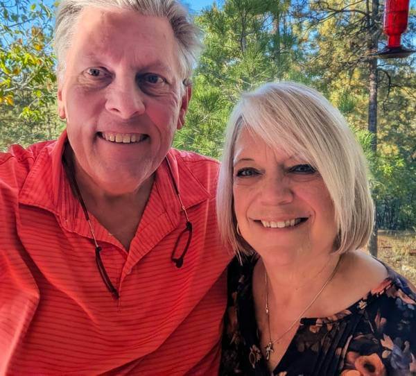 Man with gray hair and orange shirt next to lady with gray hair and dark shirt in front of trees and blue sky