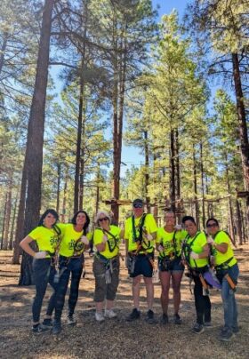 Team Sheridan, all smiles and geared up in harnesses, standing side by side at a high ropes course in Flagstaff, AZ, ready for a day of team bonding and outdoor fun.