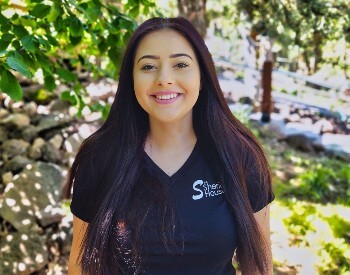 Teen girl with long black hair, in a black t-shirt, smiling and standing in front of a wooded area