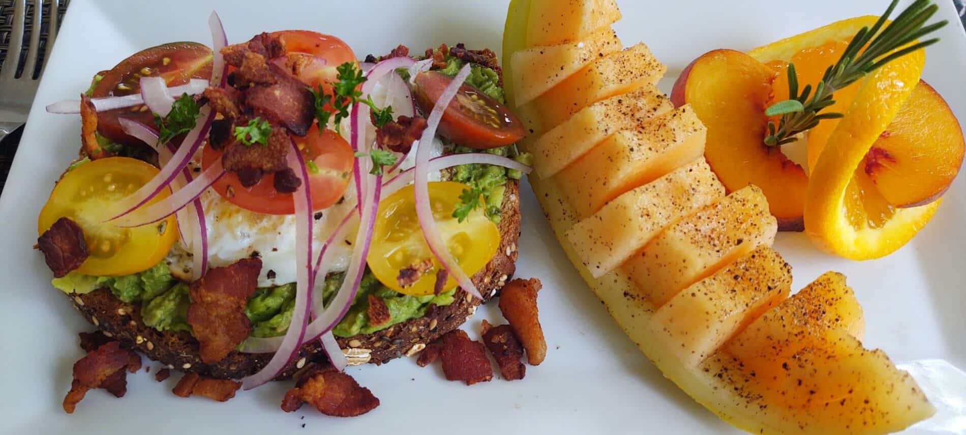 white plate with piece of toast with avocado, onion, tomato and bacon on it next to a piece of antelope and an orange spiral