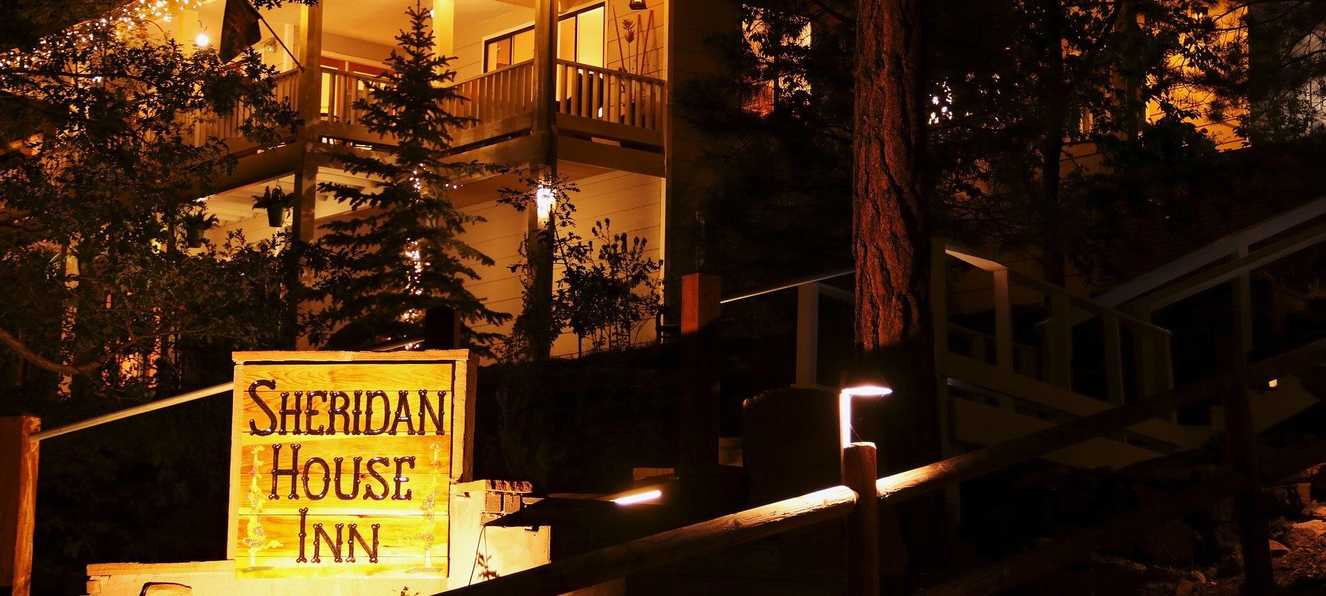 Nighttime outside of a two story home in the woods with glowing porch lights and inn sign lit up