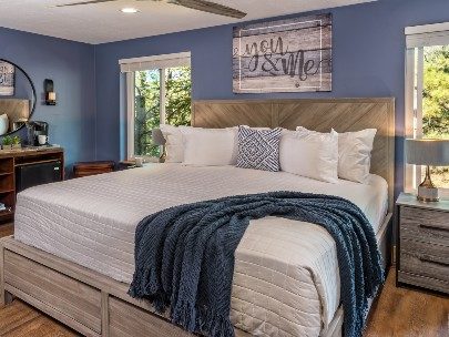 Spacious bedroom featuring a king bed with white comforter, two bright windows and dresser with circular mirror