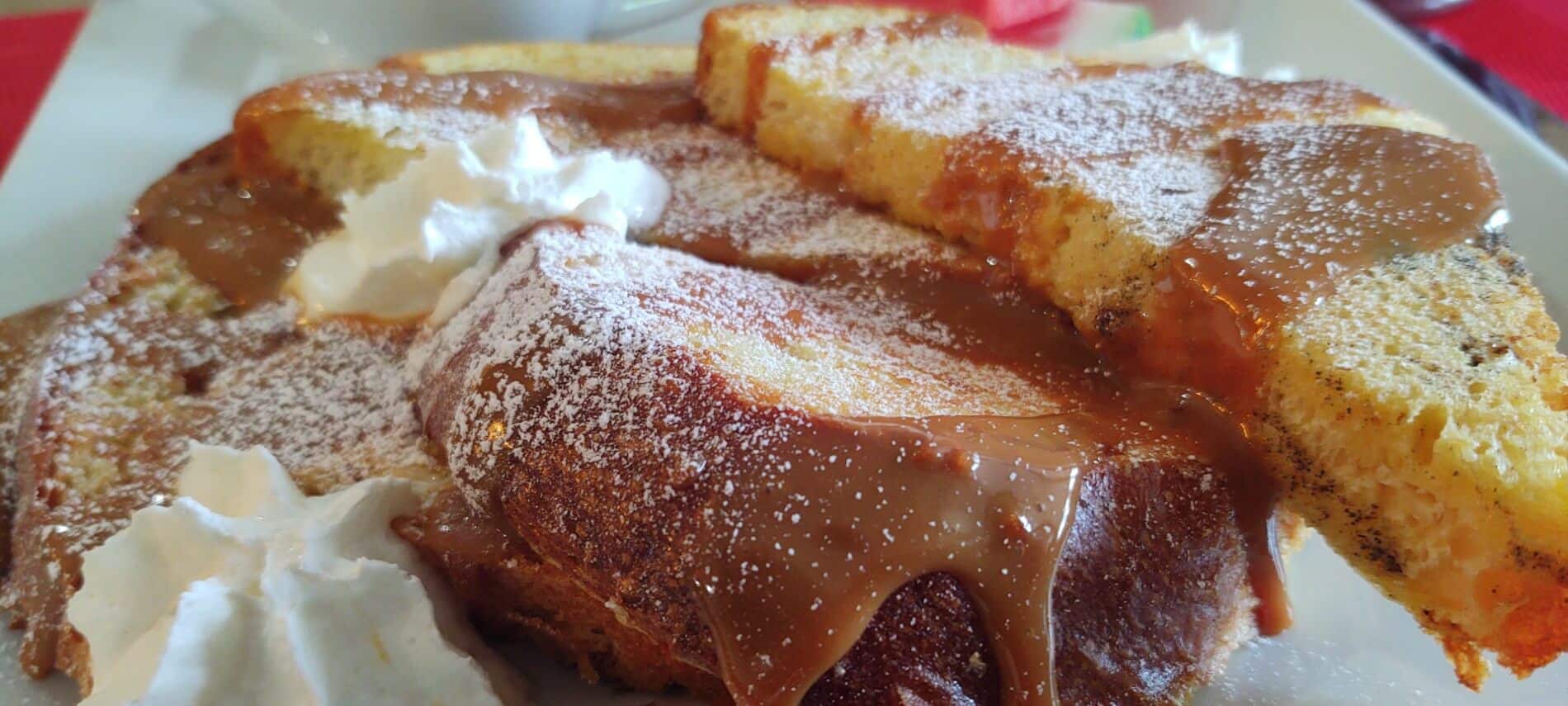 white plate with thick french toast stacked at an angle, covered with caramel sauce, whipped cream and powdered sugar