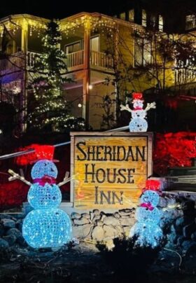 Sheridan House Inn at night during the holidays, with three illuminated snowman decorations around the sign reading 'Sheridan House Inn' and the building in the background lit up with holiday lights, including a glowing pine tree.