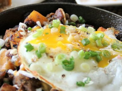 Cast iron pan filled with breakfast hash of meat and potatoes topped with fried eggs and diced green onion