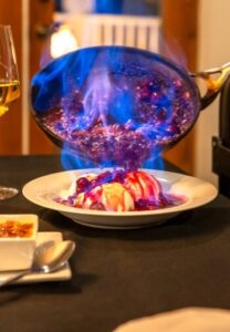 pan flaming with blue liquid being poured into a white bowl with 2 scoops of ice cream