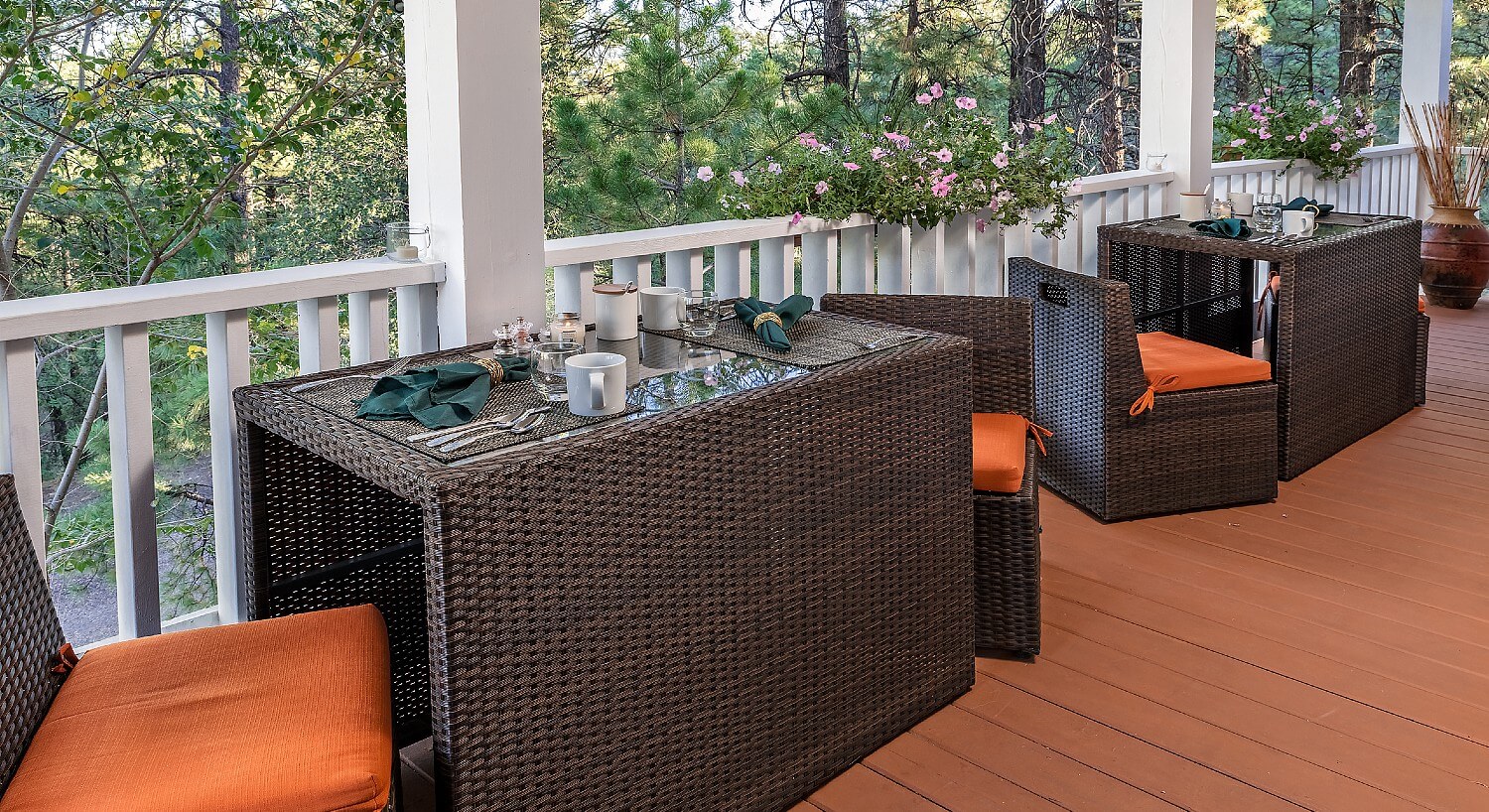 Outdoor patio with two black wicker tables and chairs set for two with flowers and lush trees in the background