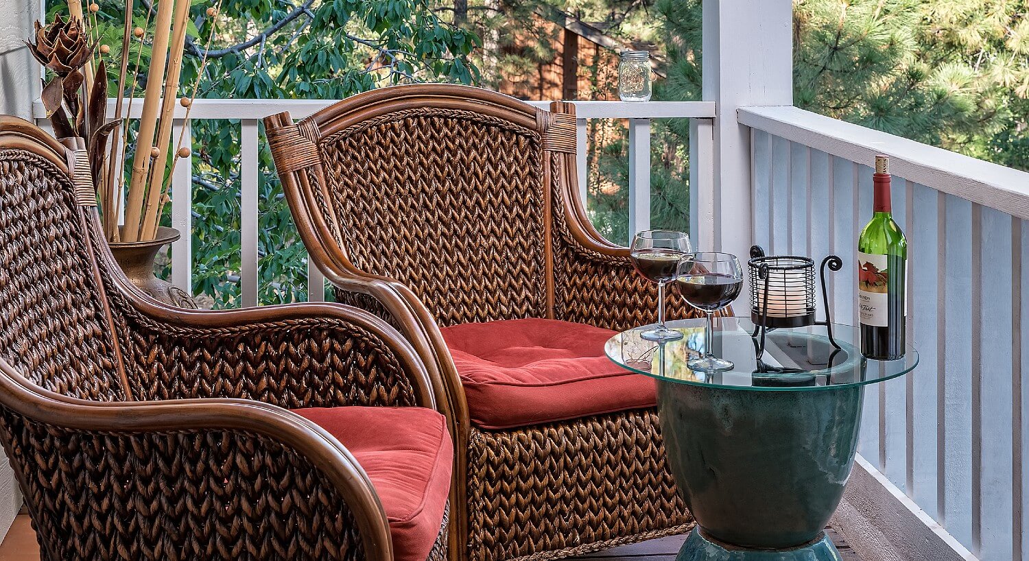 Two brown wicker chairs with red cushions outdoors on a small patio with glass table holding bottle of wine and two glasses