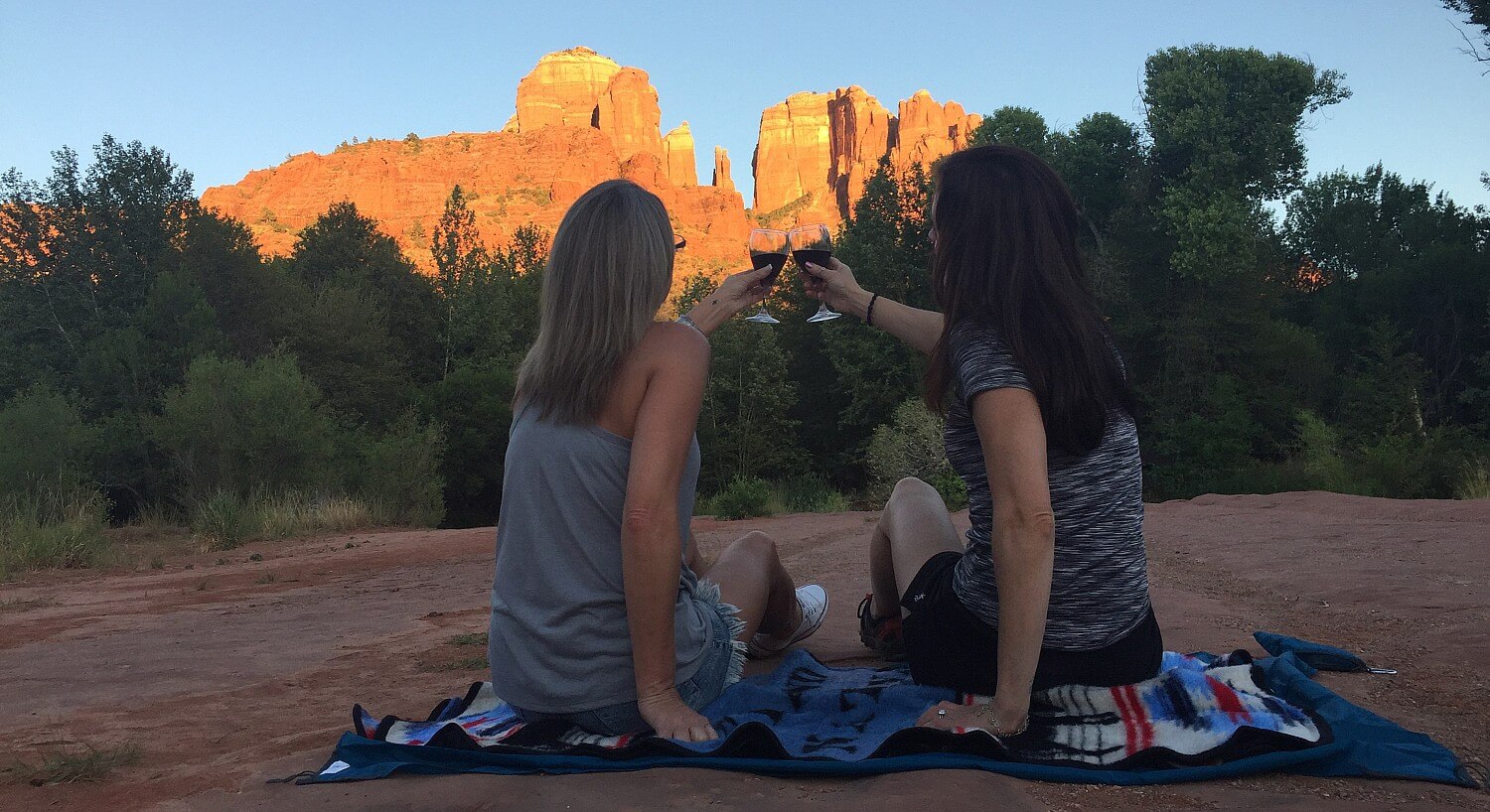 Two girls sitting on a blanket holding up wine glasses and facing tall red rocks shining in the sun