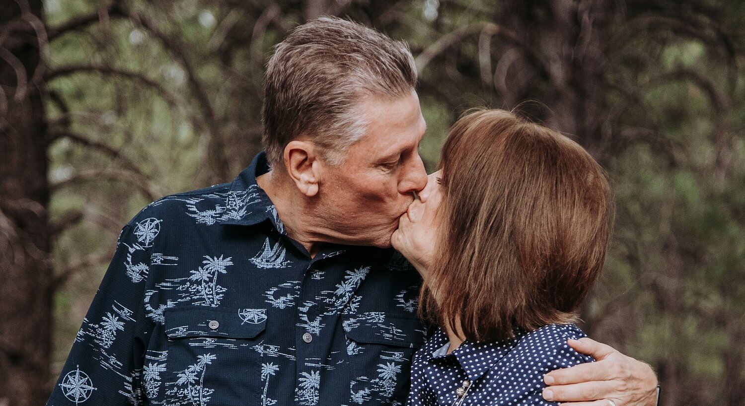 Woman with brown hair in blue and white blouse kissing a man in blue and white shirt with forest in background