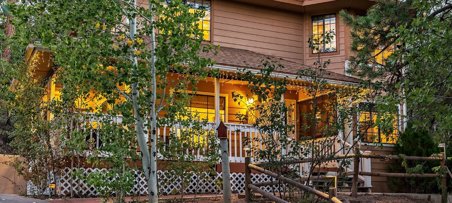 Front of a home with brown siding, many windows, and porch with white railing by birch trees