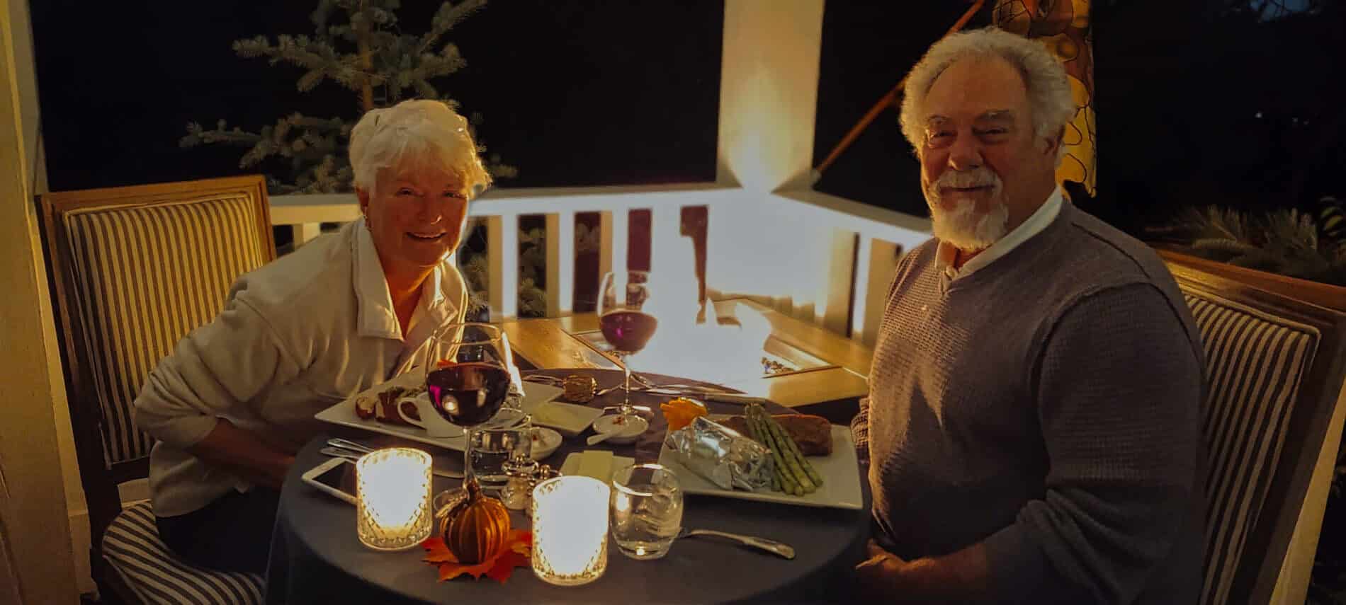 older couple sitting on balcony for dinner by candlelight with red wine glasses and fire pit burning in corner