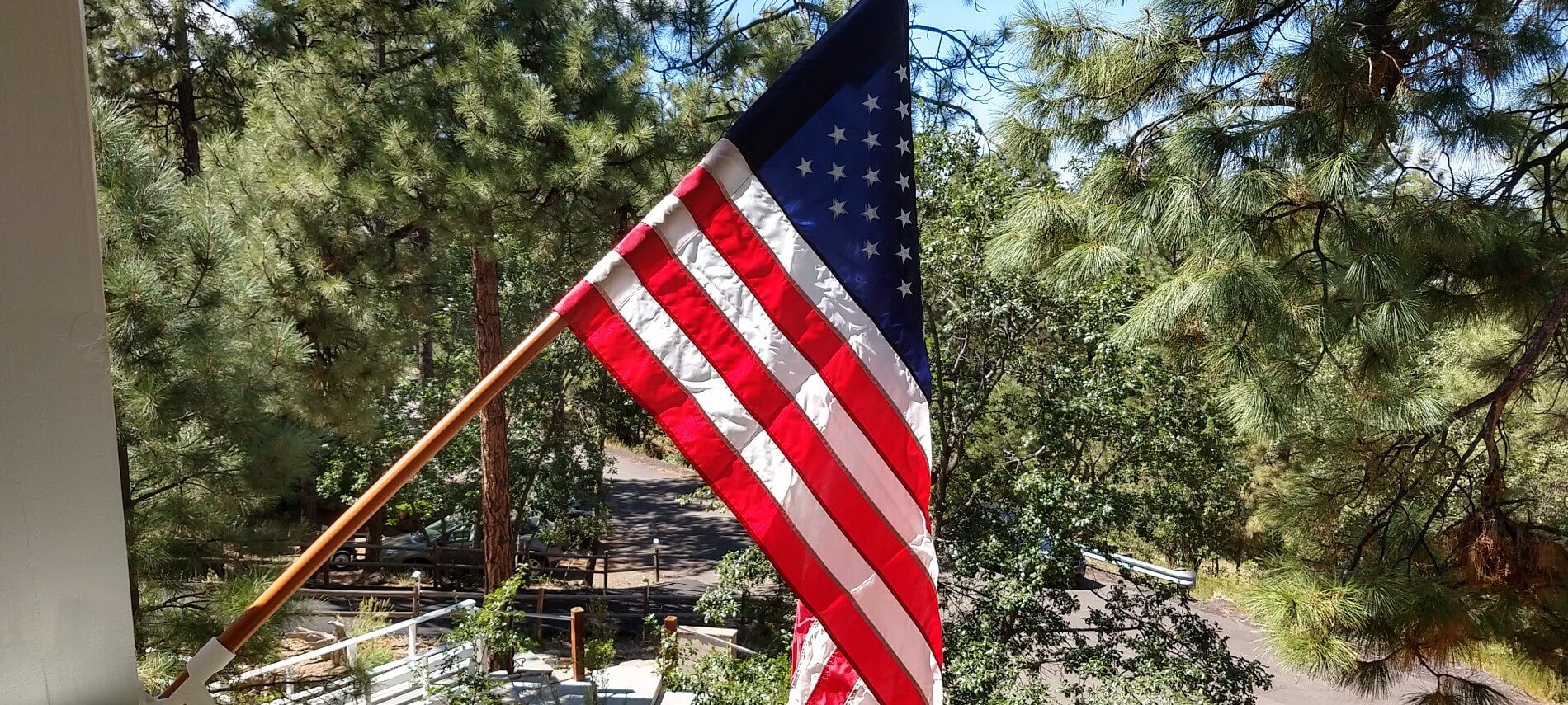 Red, white and blue American flag hanging on the side of a house with forest and driveway in the background