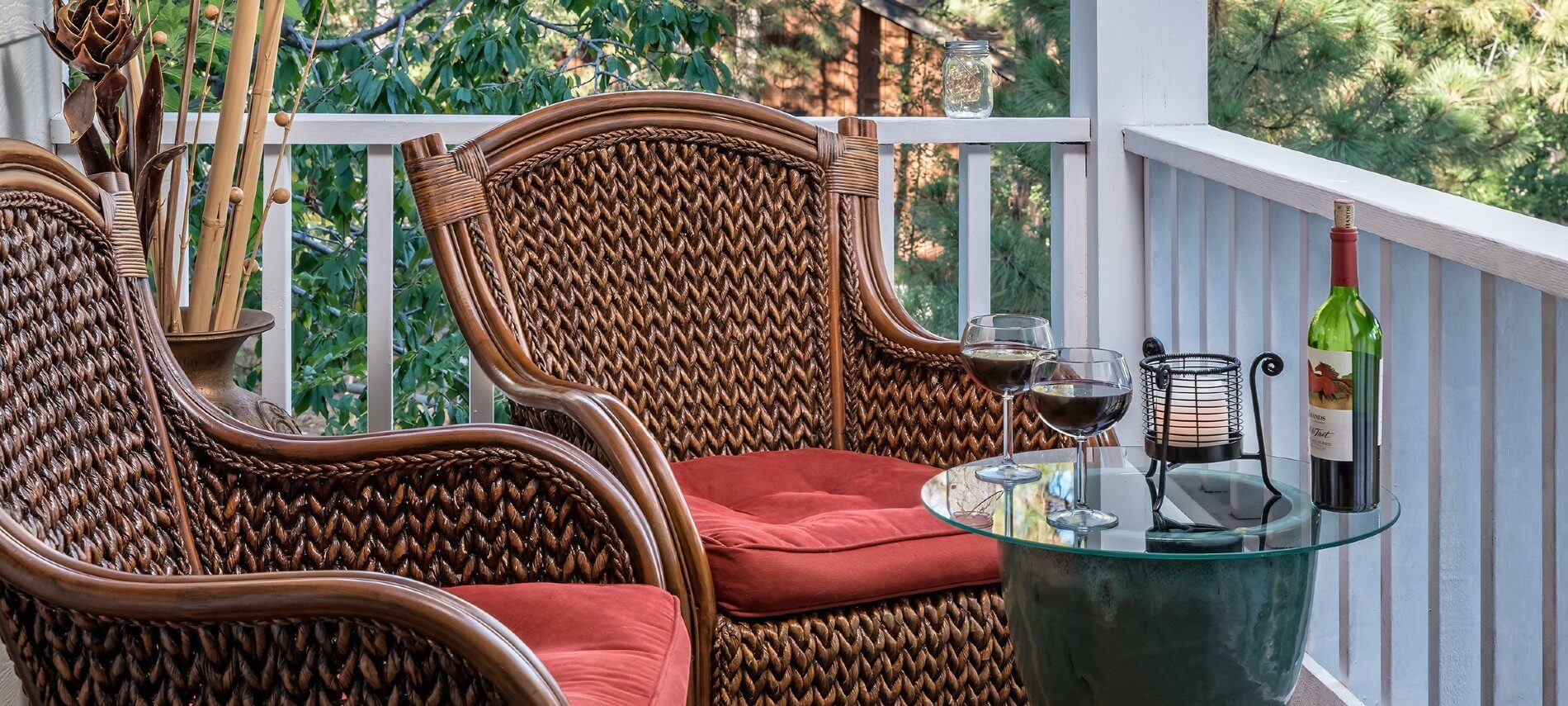 Two brown wicker chairs with red cushions on a porch with white railing and glass side table holding bottle of wine and two glasses half full