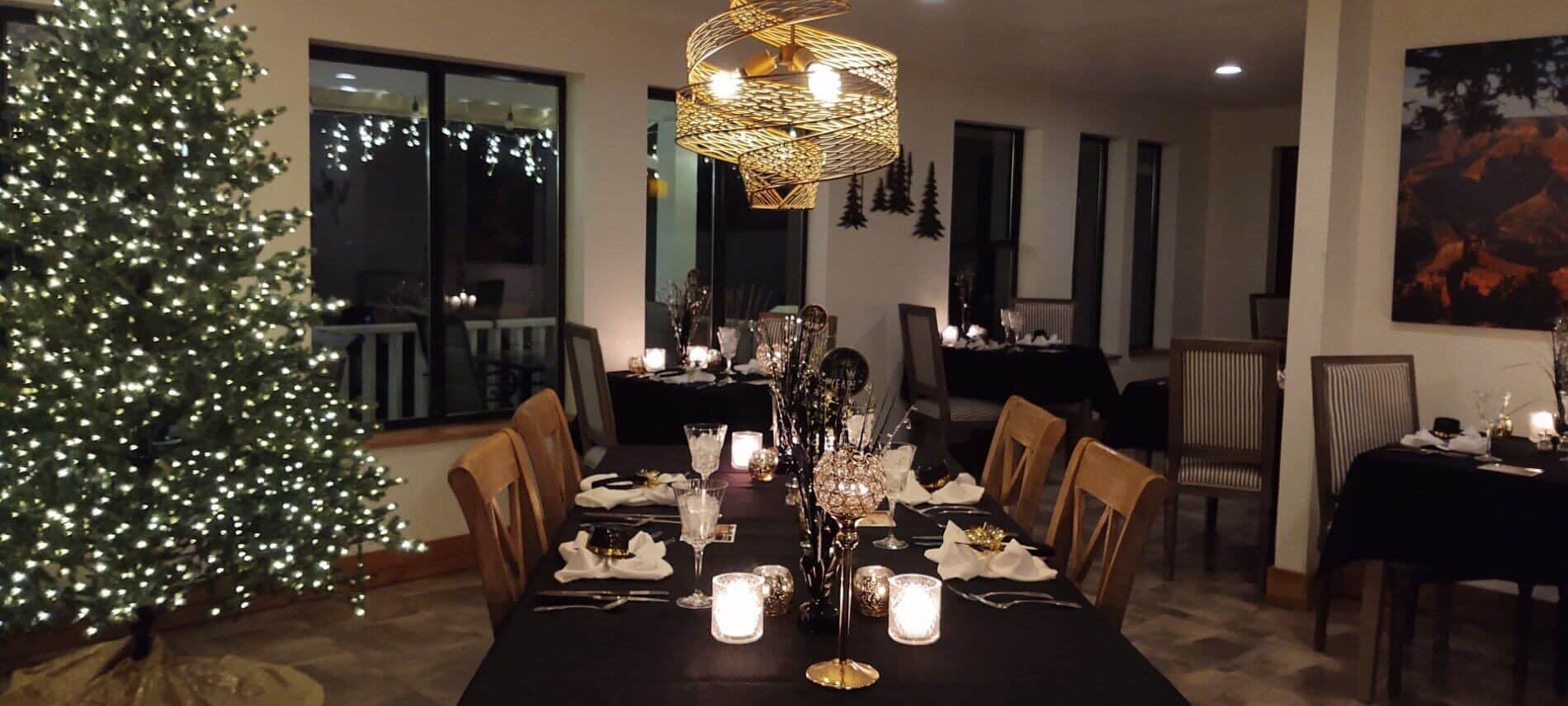 multiple tables set in dining room with white linen napkins and Christmas decorations, Christmas tree with white lights, balcony seen through window with dangling white lights