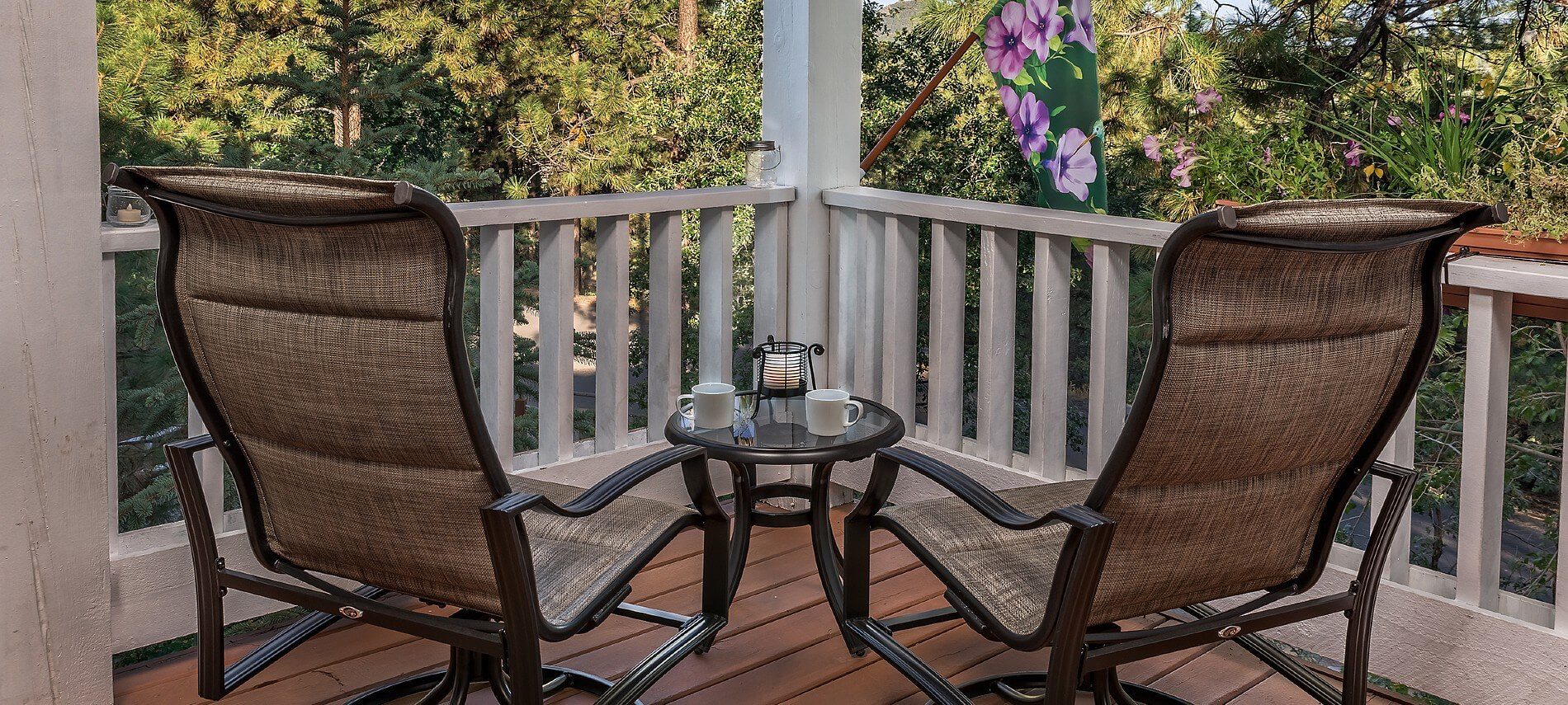 Two metal framed patio chairs with side table holding coffee cups outside viewing a wooded area