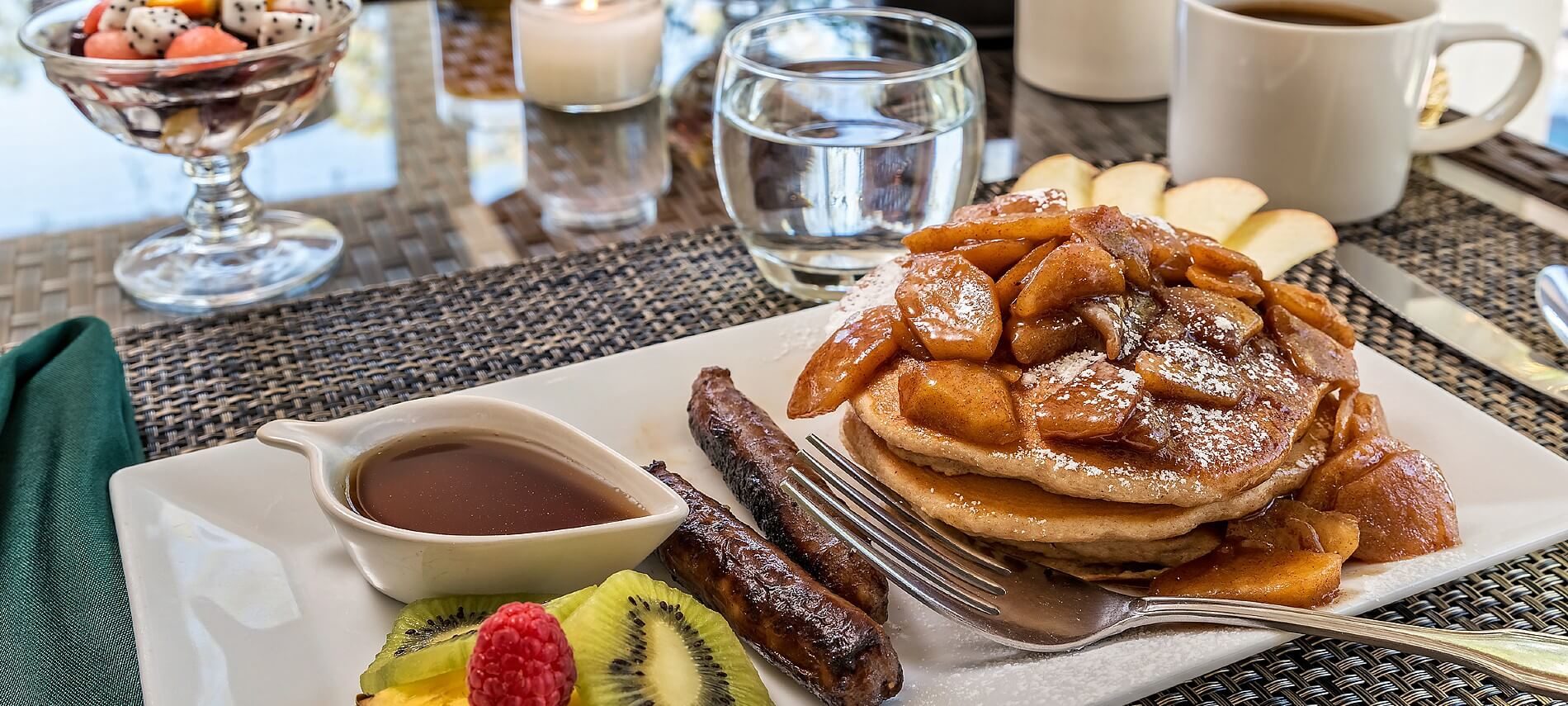 Rectangular white plate with stack of pancakes topped with fruit, two sausage links and bowl of syrup