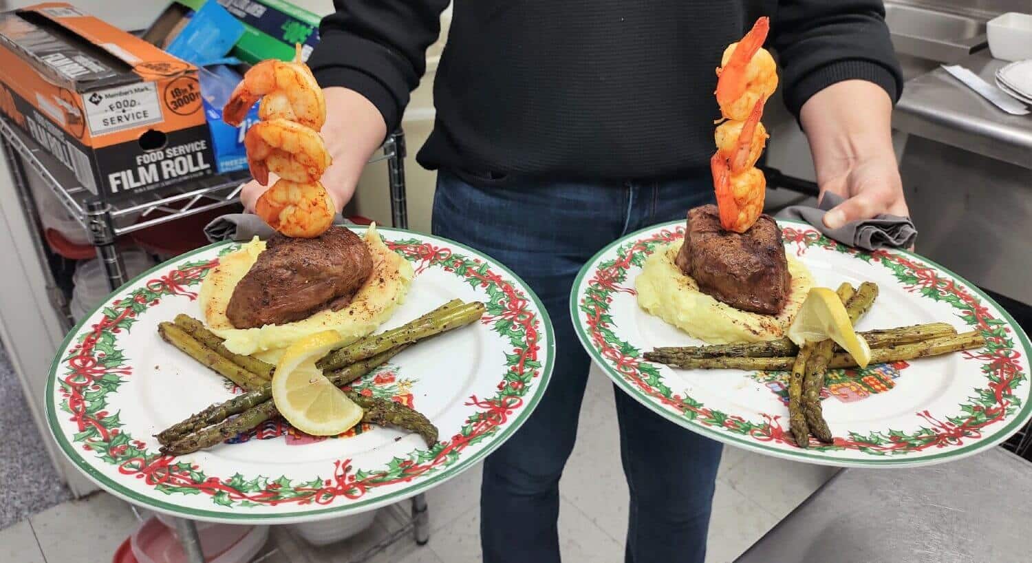 white Christmas plates being held with asparagus, an orange pepper flower, mashed potatoes, beef and shrimp on a bamboo skewer