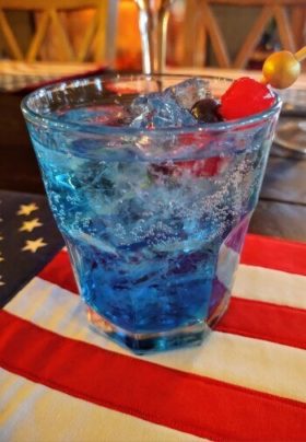 glass with a blue liquid drink and ice cubes with a cherry on a US flag placemat