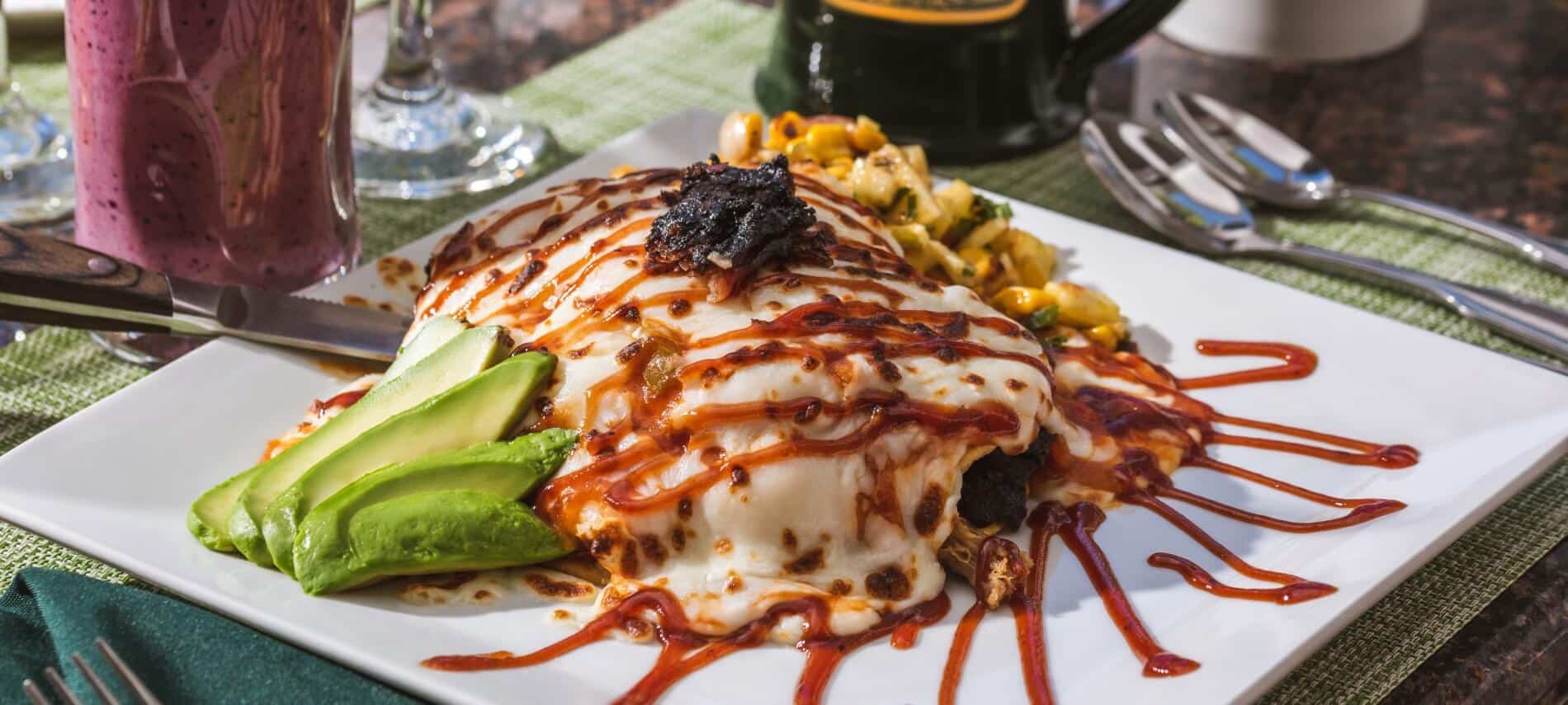 white plate with stack of stuffed tortilla shells covered in cheese with a red sauce and avocado slices and corn salsa