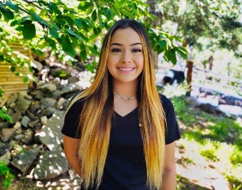 Teen girl with very long light brown highlighted hair, wearing a black t-shirt and standing in front of a wooded area