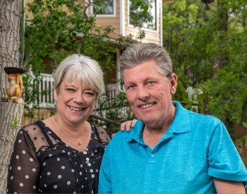 Woman with short gray hair and black and white polka dot shirt and man in slat and pepper hair with a blue shirt stand outside with a building and lots of trees behind them