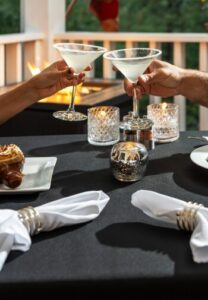 fire pit in background with a table with black tablecloth with 2 martini glasses clinking