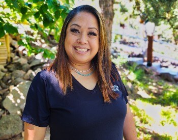 Woman with highlighted black hair, wearing a black t-shirt smiling while standing in a wooded area