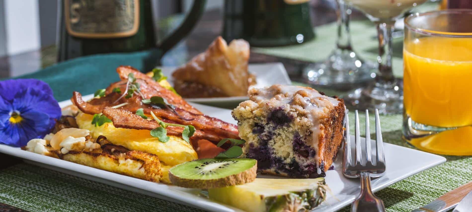 white plate with an omelet with bacon on top, a square blueberry coffee take, piece of pineapple and kiwi, with a separate plate with a triangular pastry