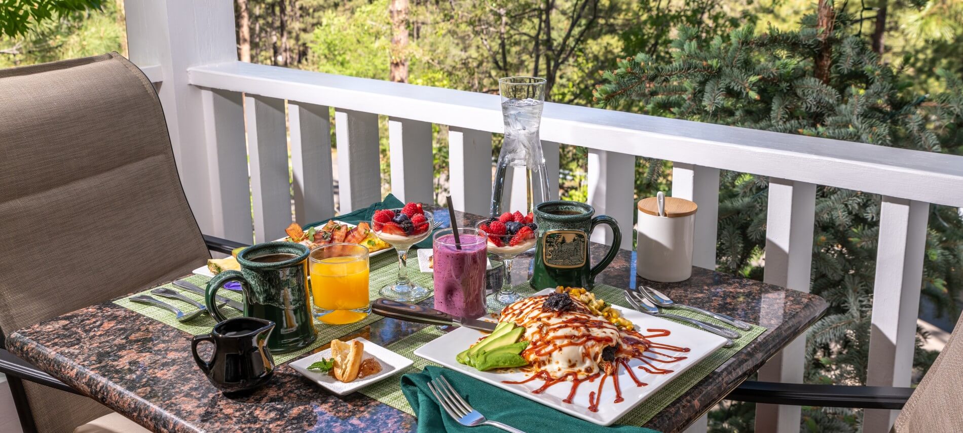 outdoor table for 2 on a porch with white railing and trees beyond it. on the square talbe are plates with food, coffee cups, juice, smoothie, little pastries and fruit