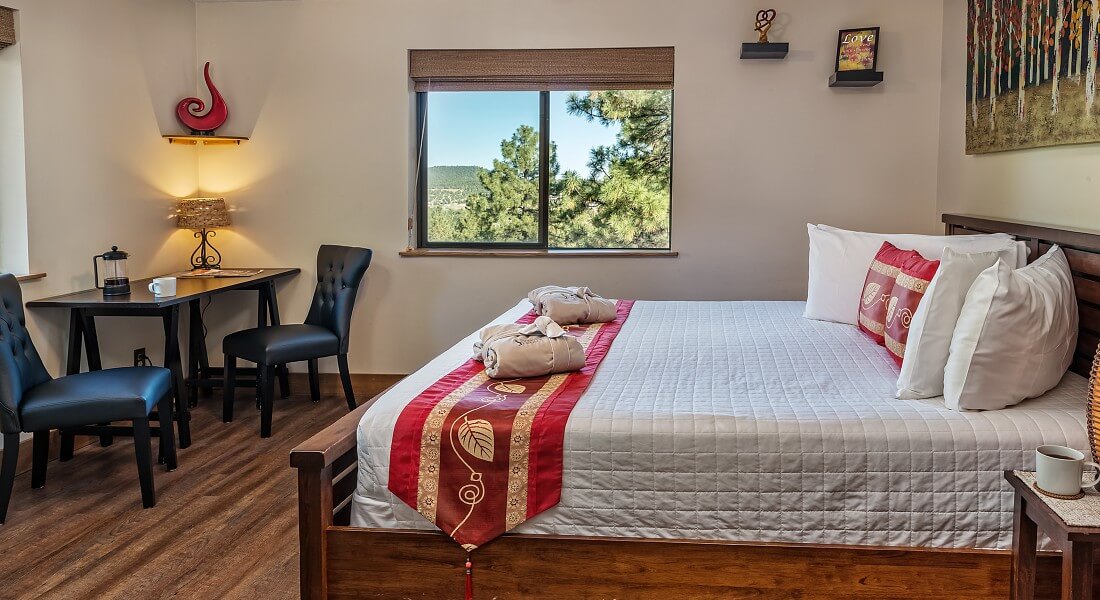 King bed with white comforter and red accent pillows in bright room with large windows and sitting desk with two black leather chairs