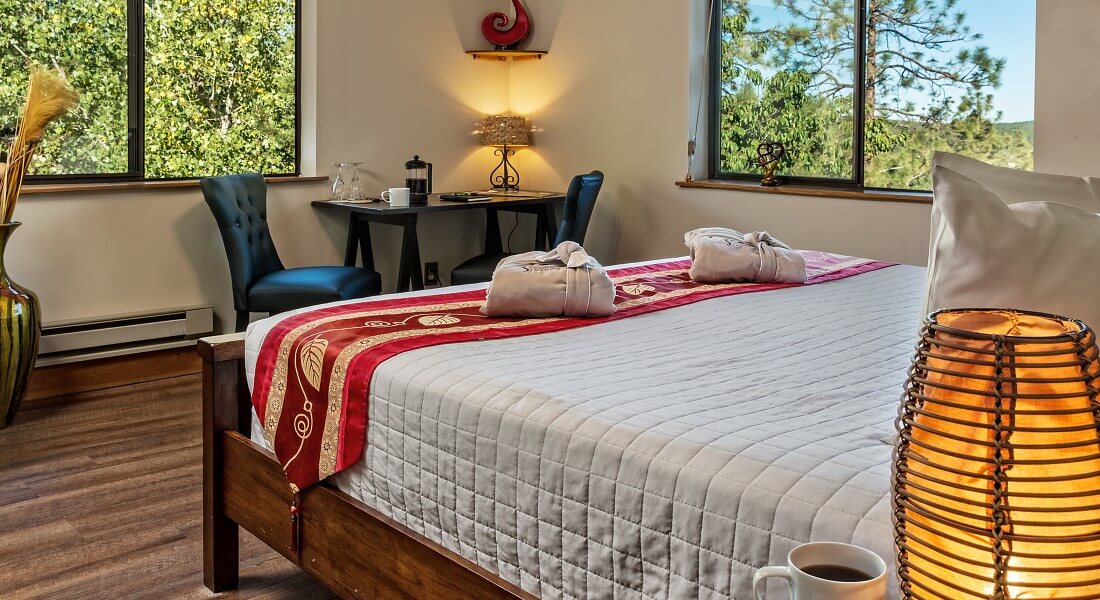 King bed with white comforter and red accents in bright room with large windows and sitting desk with two black leather chairs