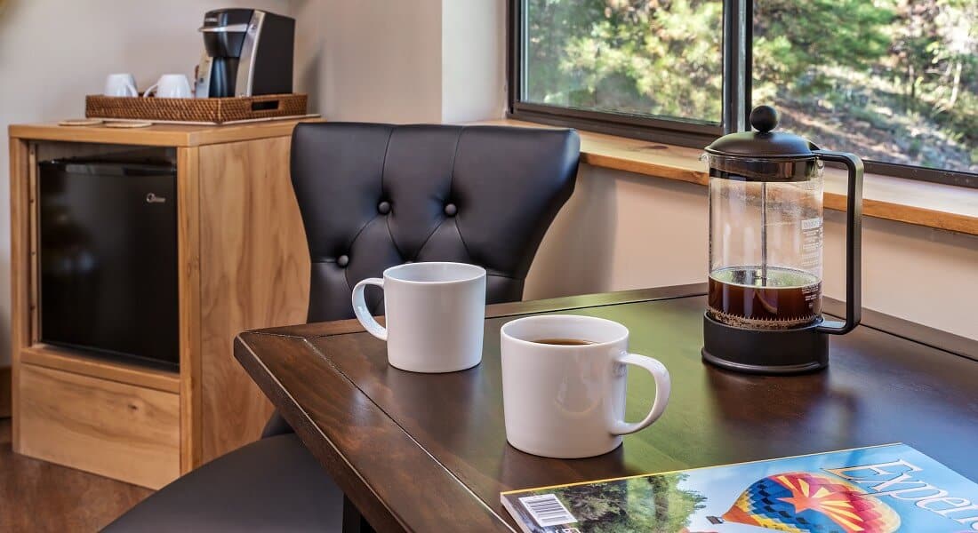 Brown table with French Press and two white coffee mugs next to a black leather chair and refrigerator cabinet