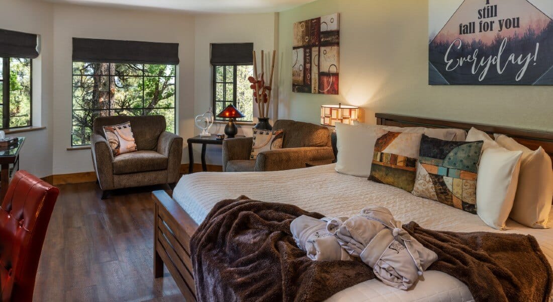 King bed with white comforter and brown accent pillows, side tables with lamps, and sitting area under three large windows