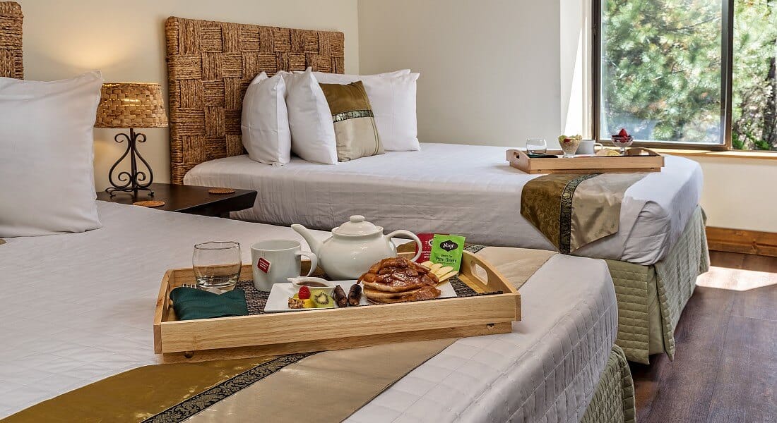 Two double beds in bright spacious room with white comforters each with a wooden tray of breakfast food and tea