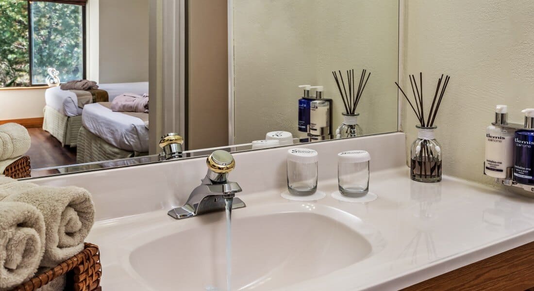 White vanity with water glasses, toiletries and rolled towels and large mirror reflecting two double beds