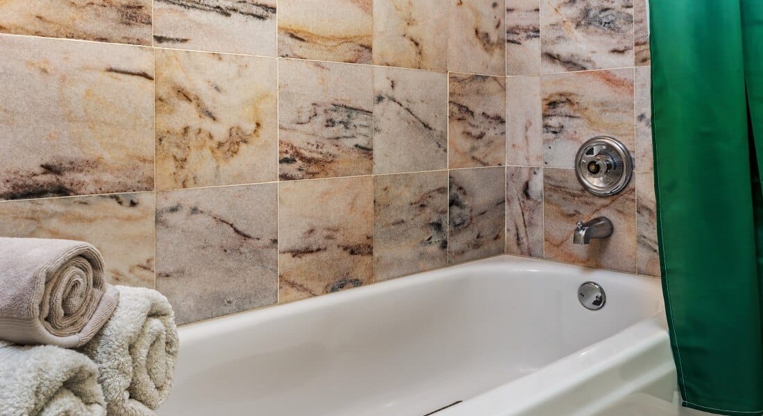 White tub and beautifully tiled shower with a green curtain and three beige rolled up towels