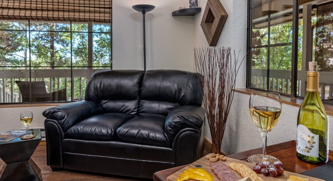 Black leather loveseat in the corner of a bright room with large windows and table with charcuterie board and wine