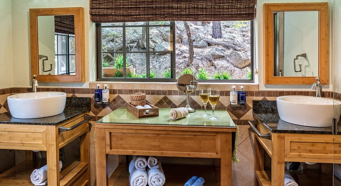 Large bathroom with three wood vanities holding rolled up towels, two round countertop sinks below mirrors and large window overlooking a forest