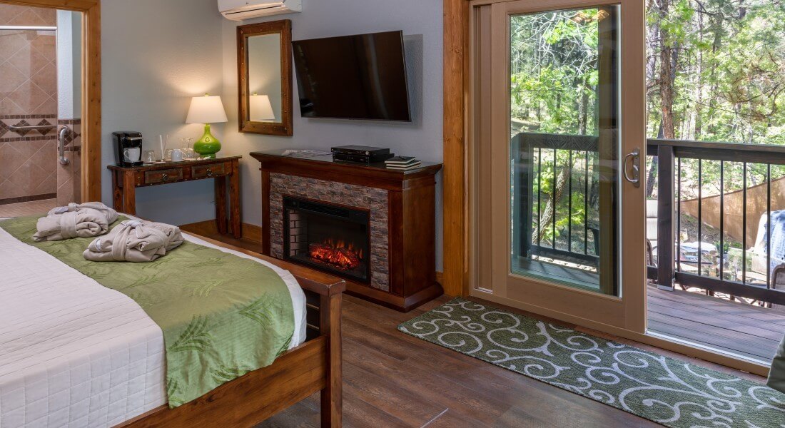 edge of a large bed facing a sliding glass door to forest, a fireplace and a large tv