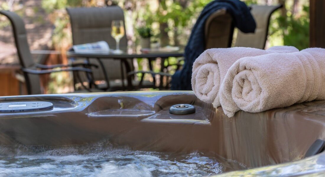 two rolled beige towels sitting on edge of bubbling hot tub with table and chairs in background by woods