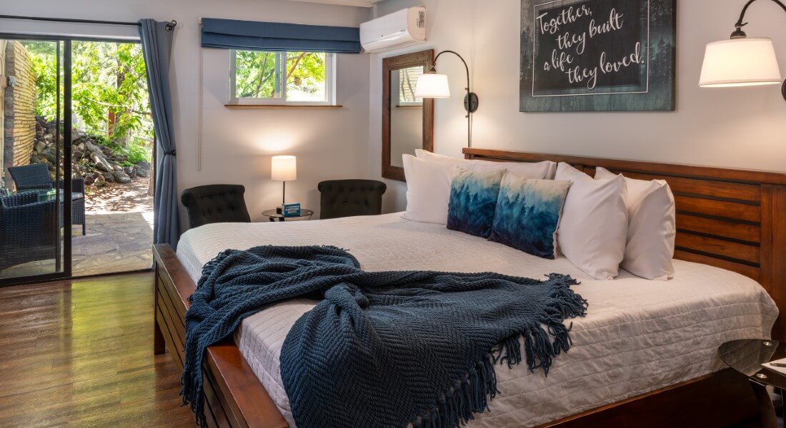 King bed with white comforter and blue accent pillows, fridge and coffee bar and wicker chair next to sliding door out to a patio