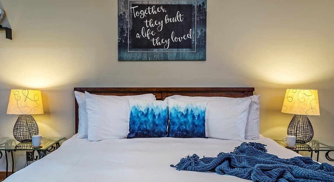 King bed with white linens and blue accent pillows with glass side tables and artwork on cream walls