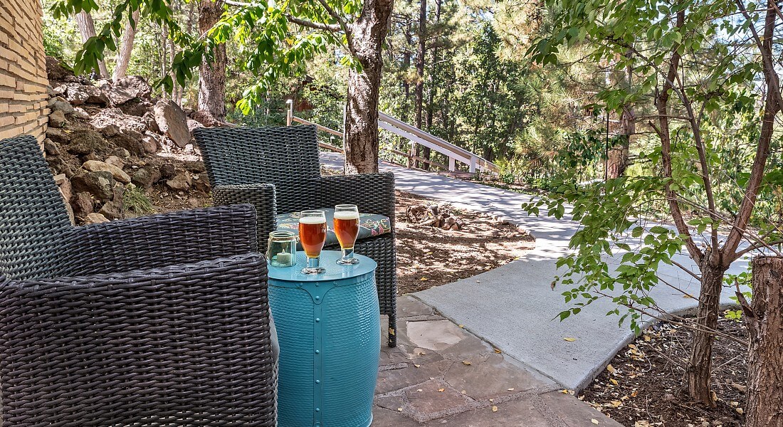 Patio with two grey wicker chairs and blue barrel table with drinks facing sidewalk into a wooded forest